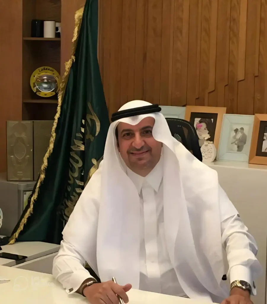 Smiling man in a white thobe and headscarf sits at a desk in a wood-paneled office with framed photos in the background.