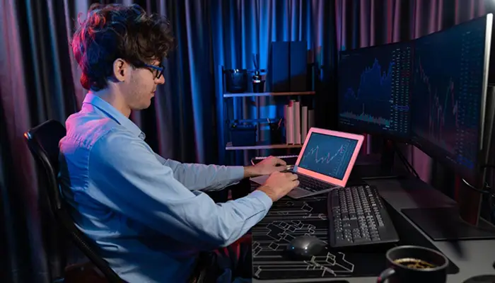 Man in a blue shirt sits at a desk with multiple monitors, working on a pink laptop displaying stock charts. Visuals show graph data on large screens behind him.