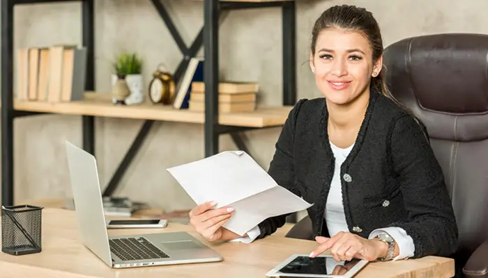 Woman in a black blazer sits at a desk in an office, holding a document with a laptop and tablet on the desk and shelves in the background.