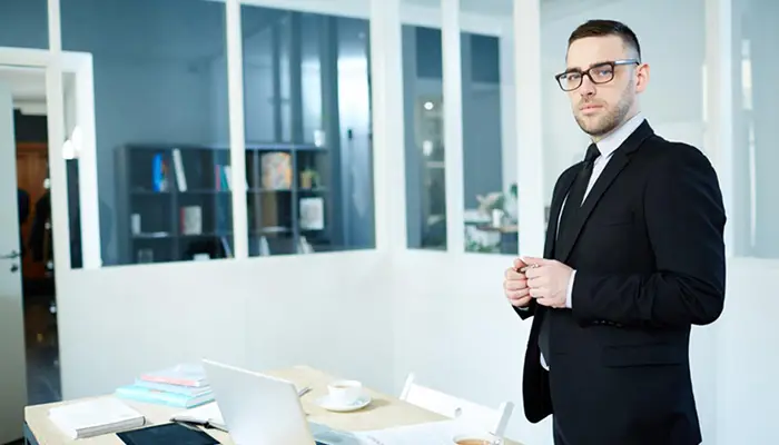 Professional man in a dark suit and glasses stands beside a desk with a laptop in a modern office conference area.