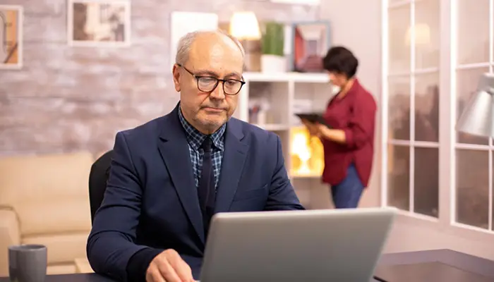 Senior man in a navy suit typing on a laptop at a desk, with a woman reading a tablet in the background.