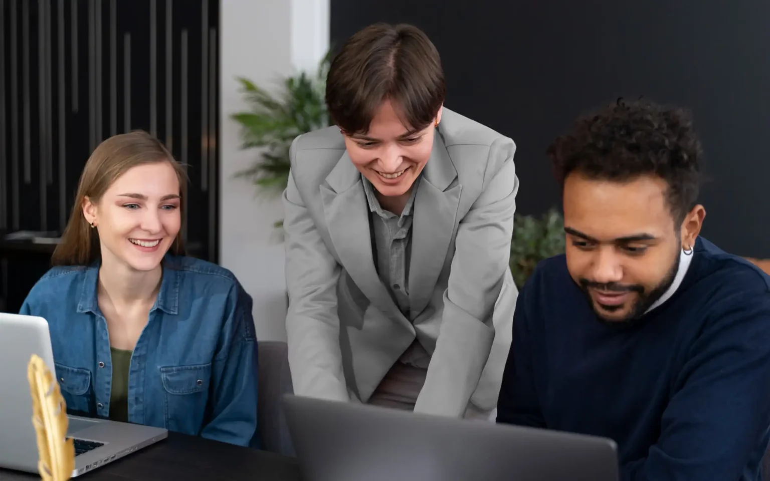 Three coworkers collaborating around a laptop, with the woman in a denim shirt smiling on the left and a man in a dark sweater on the right.