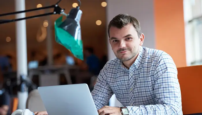 Man in a checkered shirt working on a silver laptop at a bright coworking space.