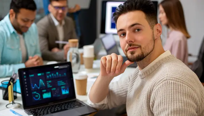 Man in a beige sweater at a laptop showing charts, in a busy office with coworkers and coffee cups nearby.